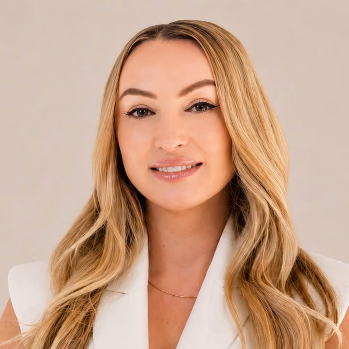 Blonde woman with long wavy hair wearing white sleeveless blazer, smiling slightly against a beige background.