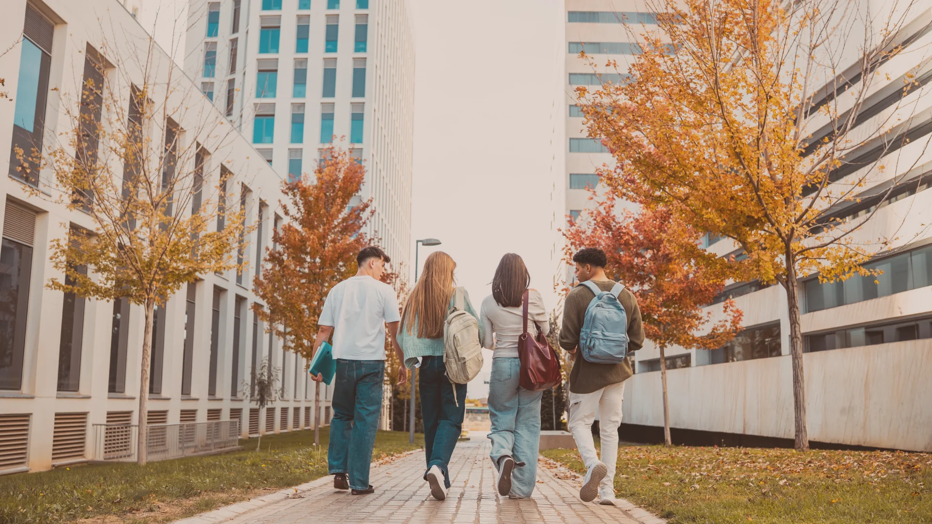 Four students seen from behind walking along a brick path between modern white university buildings, surrounded by trees with orange and yellow autumn leaves.