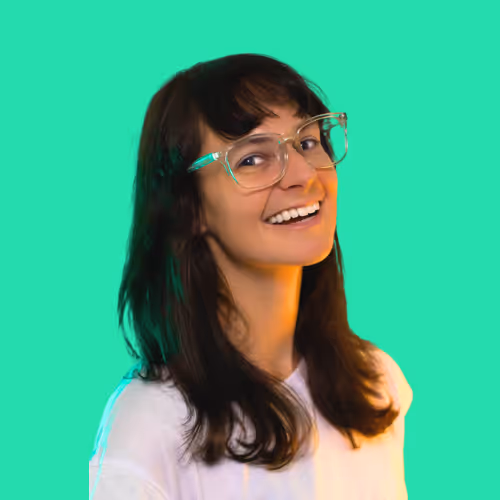 Smiling woman with long dark hair and clear glasses wearing a white shirt against a bright turquoise background.