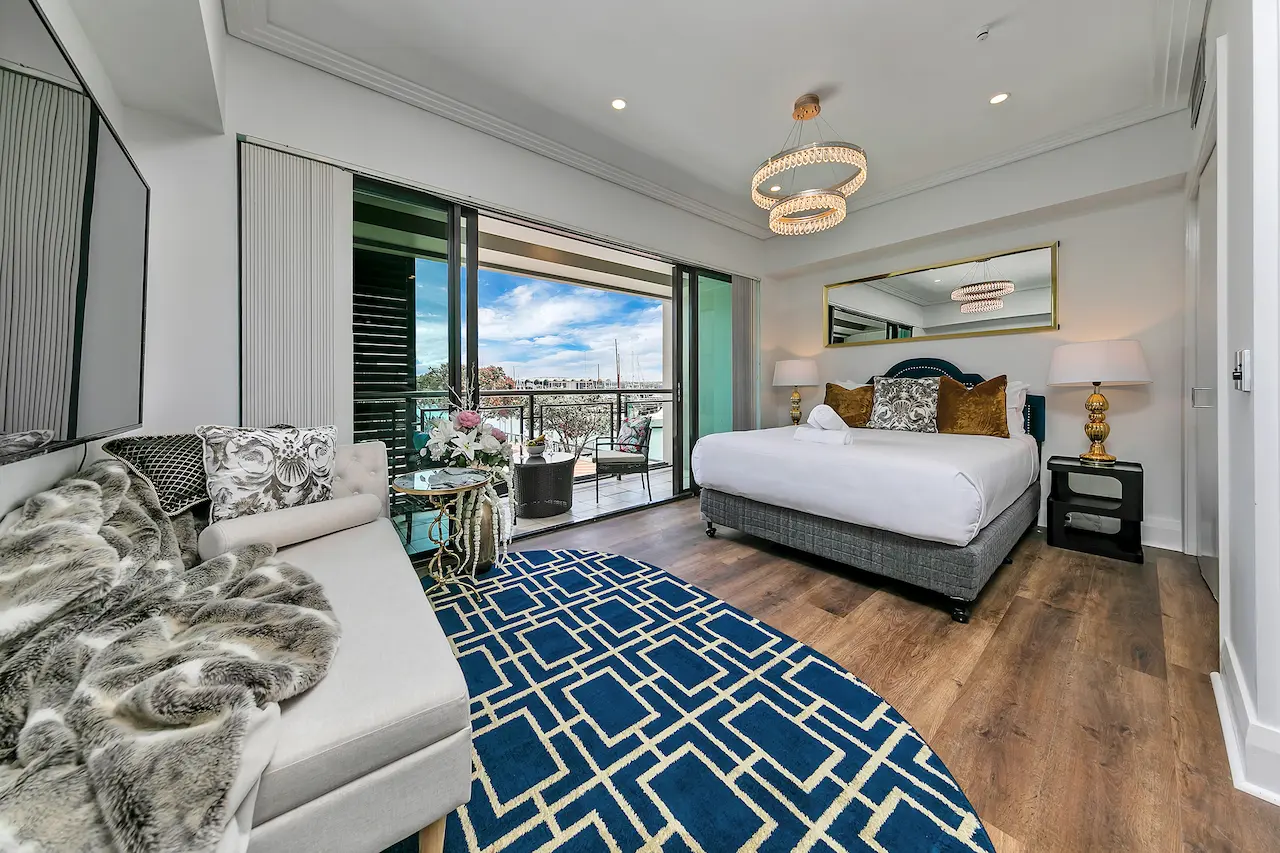Modern bedroom with a large bed, two bedside lamps, a blue geometric rug, a sofa with pillows and fur blanket, and sliding glass doors opening to a balcony with chairs and a view of the waterfront.