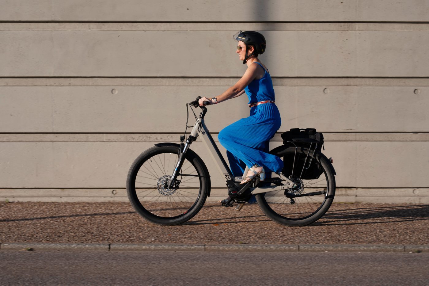 A woman in a blue dress riding a Moustache Mardi 27 commuter e-bike along a city street