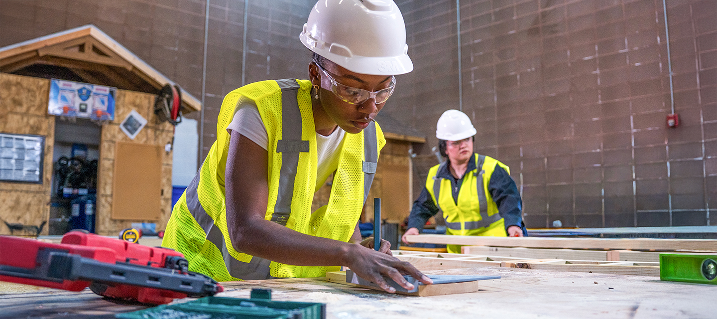 Two construction workers in a workshop wearing white hard hats, safety glasses, and yellow reflective vests; one is drawing or measuring on a wooden board with a square tool while the other stands behind with wooden planks.