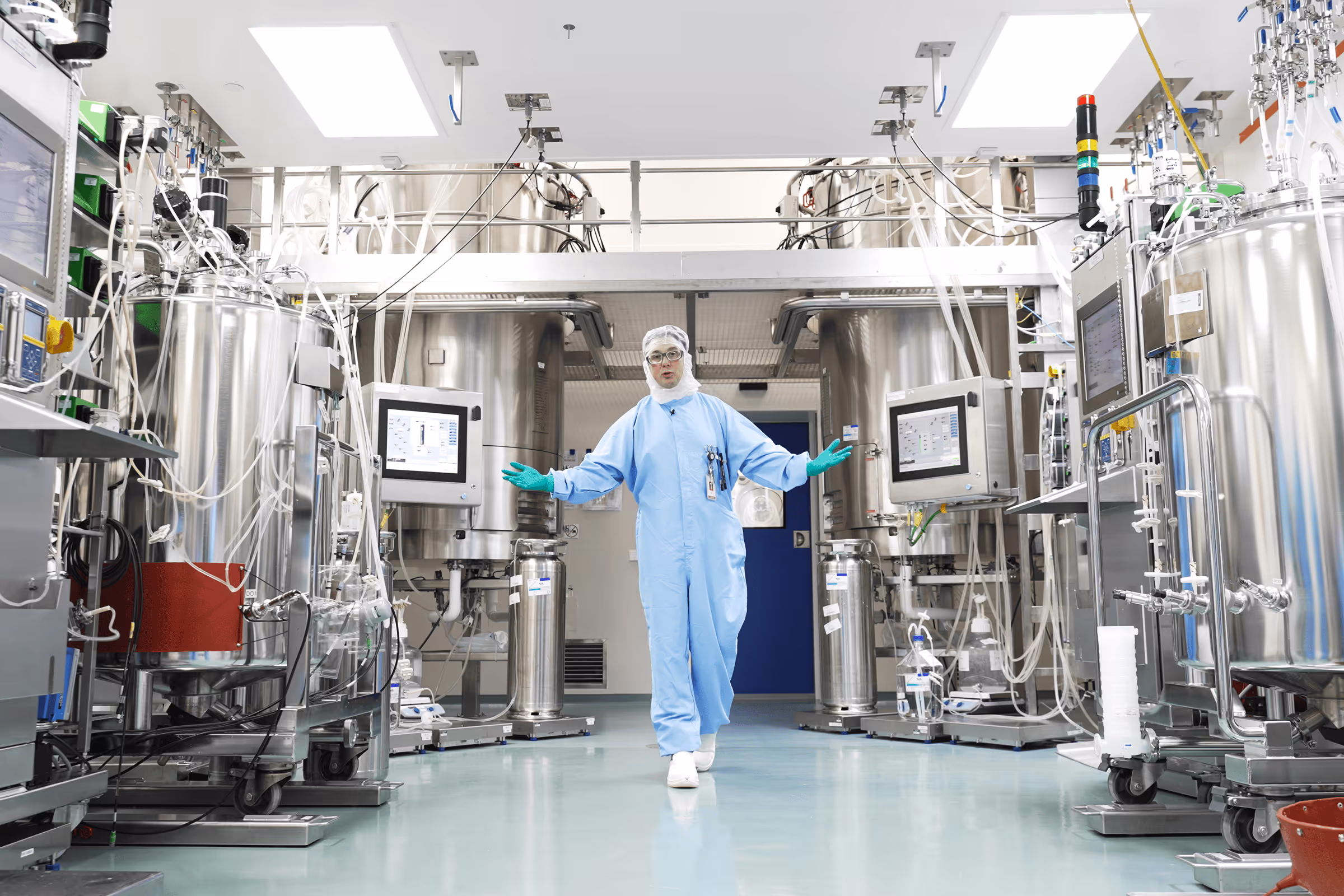 Lab technician in blue protective suit and gloves standing in a clean pharmaceutical manufacturing facility with stainless steel bioreactors and control monitors.