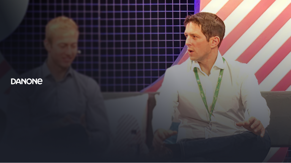 Smiling woman with long brown hair sitting against a textured dark wall with Danone logo on the left.