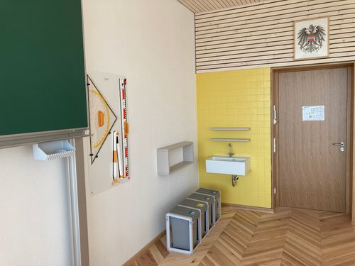 Klassenraum mit Tafel, einem großen Geodreieck an der Wand und einem Handwaschbecken
