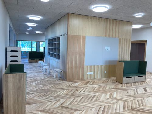 Innenraum mit Holzfußboden, Regalen, grüner Sitzbank und weißen Hockern.