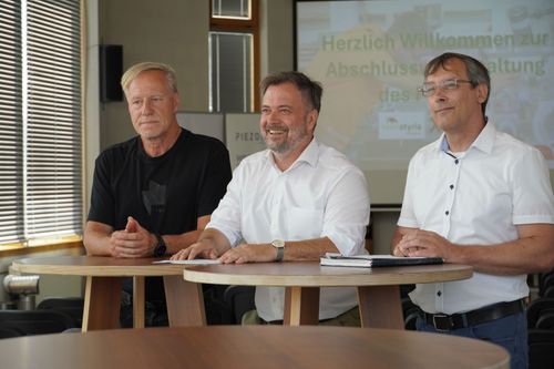 Drei Männer sitzen an einem runden Holztisch in einem modernen Raum mit Fensterjalousien, im Hintergrund eine Präsentationsfolie mit dem Text 'Herzlich Willkommen zur Abschlussveranstaltung'