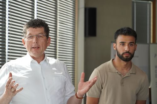 Zwei Männer stehen nebeneinander in einem Raum mit Jalousien, der linke Mann trägt ein weißes Hemd und gestikuliert mit den Händen, der rechte Mann trägt ein beiges Poloshirt.