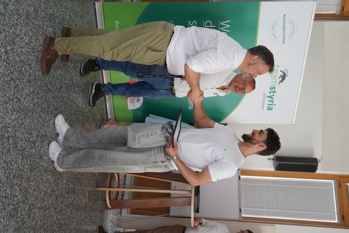 Zwei Männer geben sich die Hand, einer trägt weiße Hemd und beige Hose, der andere weißes T-Shirt und graue Jeans, im Hintergrund Banner mit Team Styria Logo
