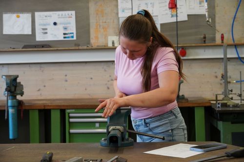 Junge Frau mit rosa T-Shirt und blauer Jeans arbeitet an einer Werkbank mit Schraubstock in einer Werkstatt