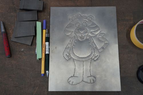 Metallplatte mit ausgestanztem Umriss eines lachenden Tigers, daneben Stifte und Schleifpapier auf Werkbank