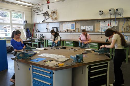 Vier junge Frauen arbeiten an Werkbänken in einer Werkstatt mit Werkzeugen und technischen Zeichnungen