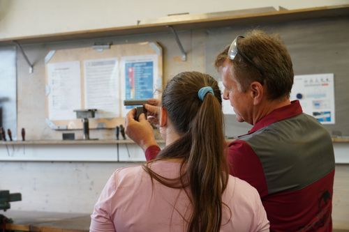 Zwei Personen in Werkstatt beim Vermessen eines Metallteils mit Messschieber, eine trägt rosa Hemd, andere rote Arbeitskleidung