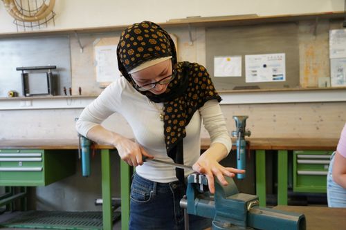 Frau mit Kopftuch und weißem Langarmshirt feilt mit einer Feile an einem Metallstück, das in einem Schraubstock in einer Werkstatt eingespannt ist