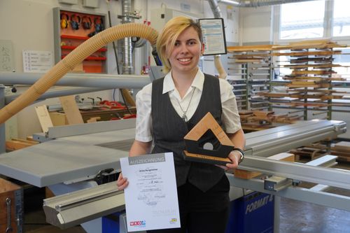 Lehrling Anna steht in einer Werkstatt und hält eine Auszeichnung und eine hölzerne Trophäe in Form eines Dreiecks