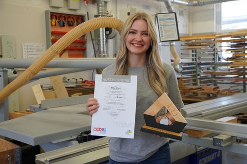 Lehrling Stina hält eine Auszeichnung und einen Holzpreis in einer Werkstatt mit Maschinen und Werkzeugen