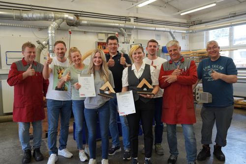 Gruppe von zehn Personen in Werkstatt, einige halten Urkunden und Auszeichnungen, mehrere tragen rote Arbeitskittel, Daumen hoch Geste