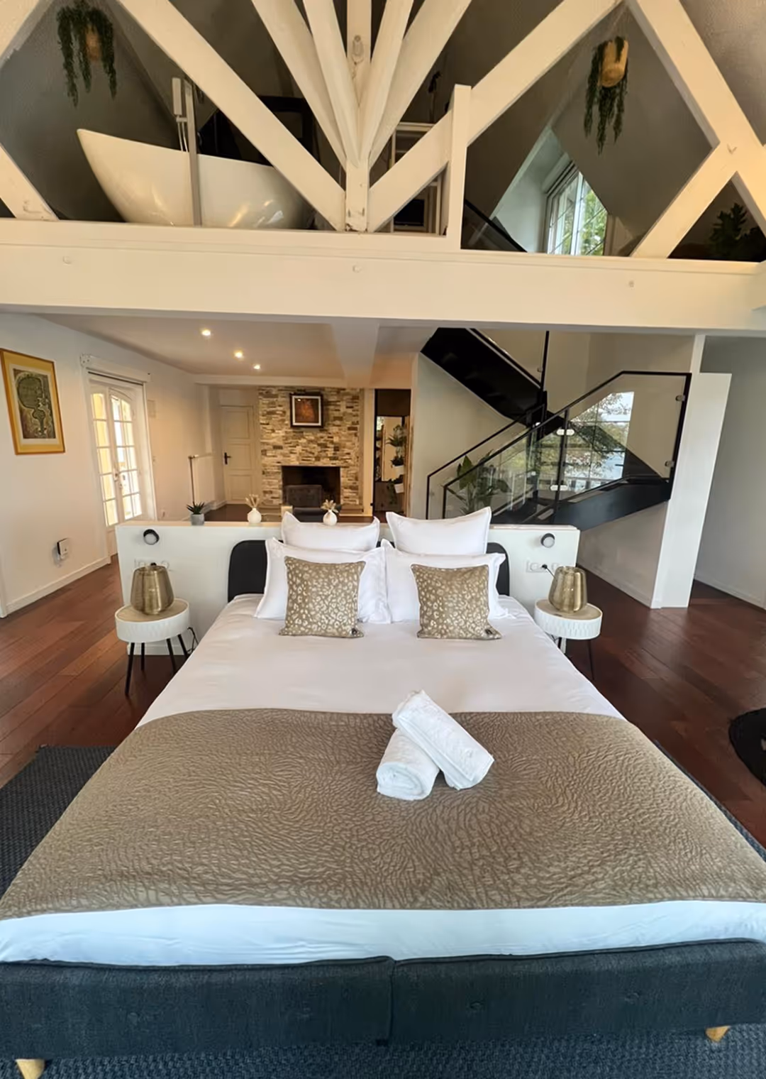 Modern bedroom with a large bed featuring white and gold pillows, beige blanket, and rolled towels, with a fireplace and staircase in the background.