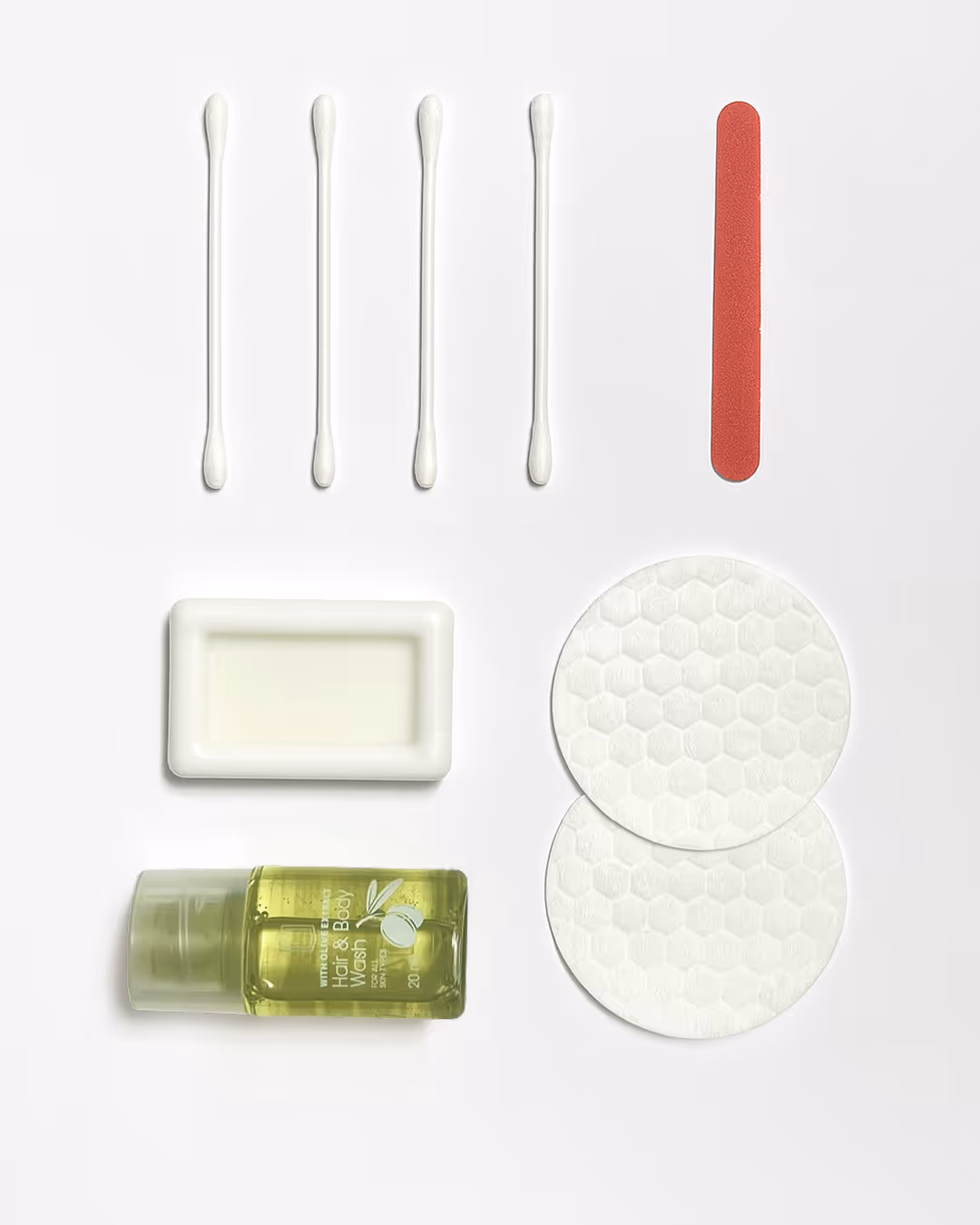 Four cotton swabs, a red nail file, a bar of soap in a white dish, two round cotton pads with hexagonal patterns, and a small bottle of lime-scented hand and body wash arranged on a white background.