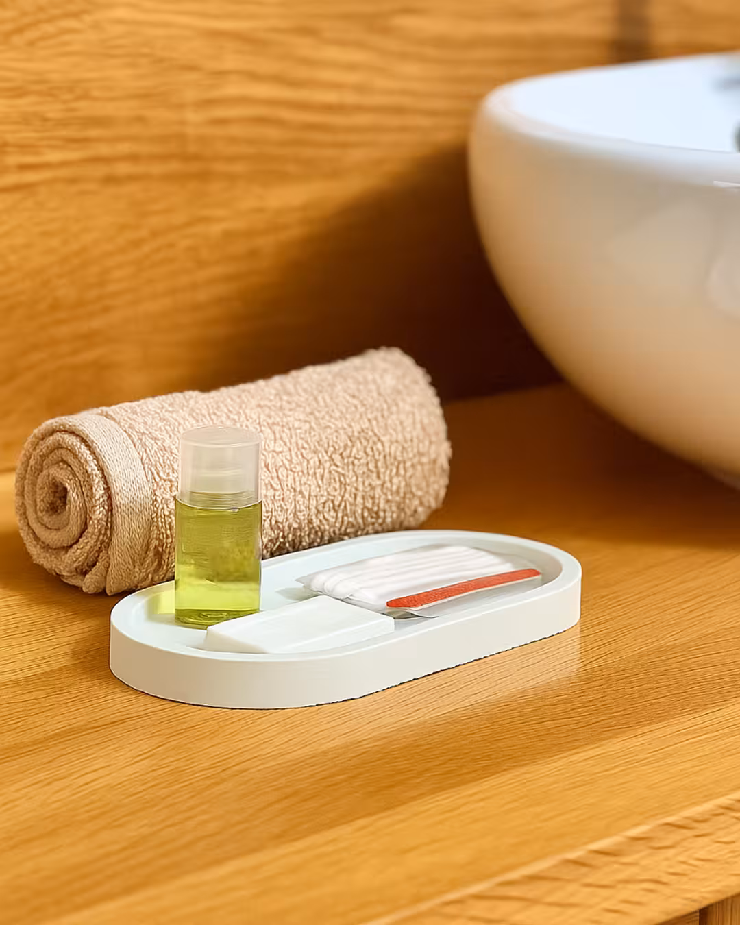 Rolled beige towel and white tray holding small toiletry items including a green liquid bottle, soap, cotton pads, and nail file on wooden bathroom counter.