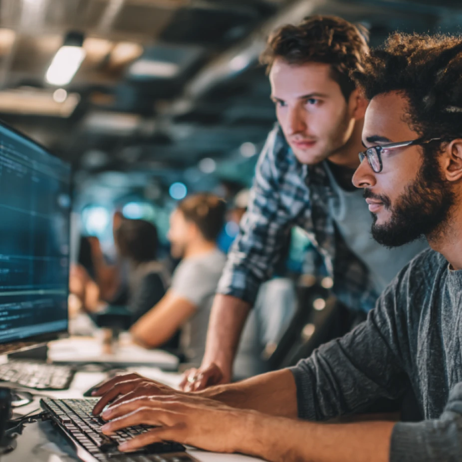 Two team members at an AI-powered SEO agency collaborate intently in a modern workspace, analyzing and coding on a dual-monitor setup filled with technical data and search engine optimization tools.