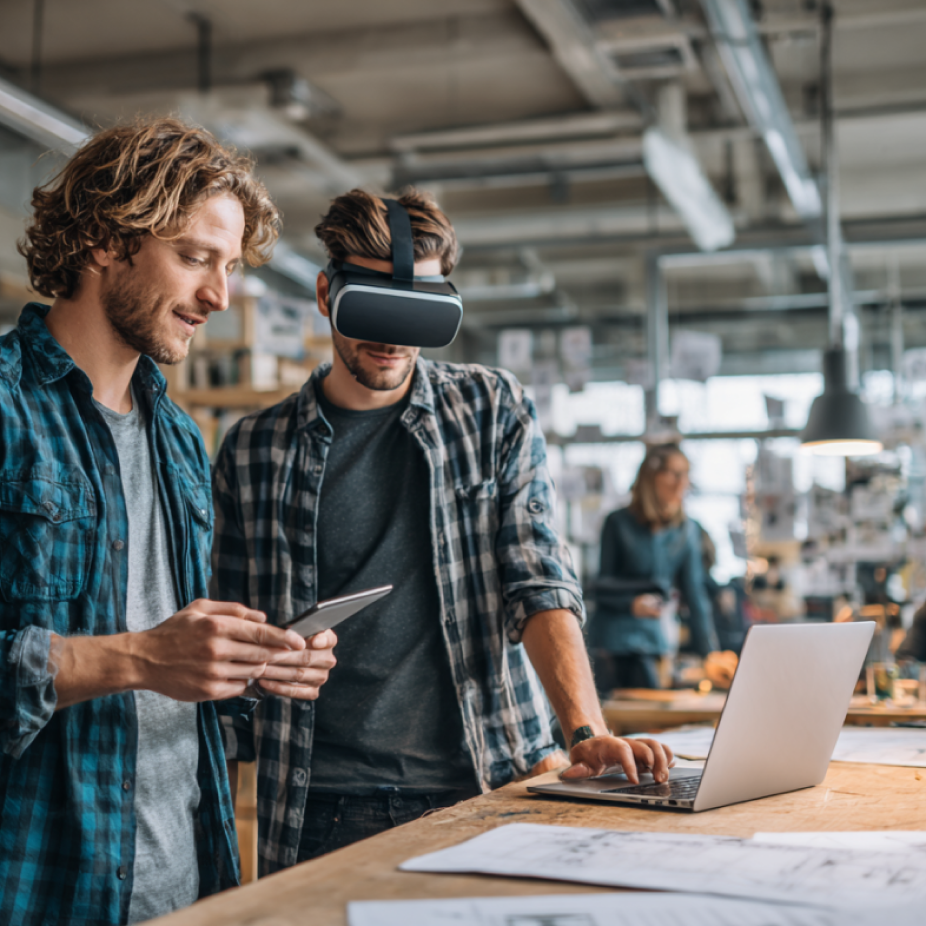 Two casually dressed creative professionals collaborate in a design studio, with one using a VR headset and the other holding a tablet. They stand beside a laptop and design sketches spread across a large wooden table. The dynamic workspace and use of technology suggest a brainstorming session focused on immersive brand design or product development.