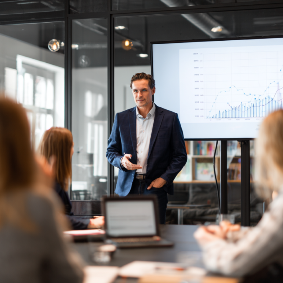 A confident businessman in a navy suit presents a B2B branding strategy to a team in a modern office meeting room. Behind him, a screen displays a detailed line and bar graph showing business growth trends. Team members seated at the table are attentively listening, with laptops and notes in front of them.