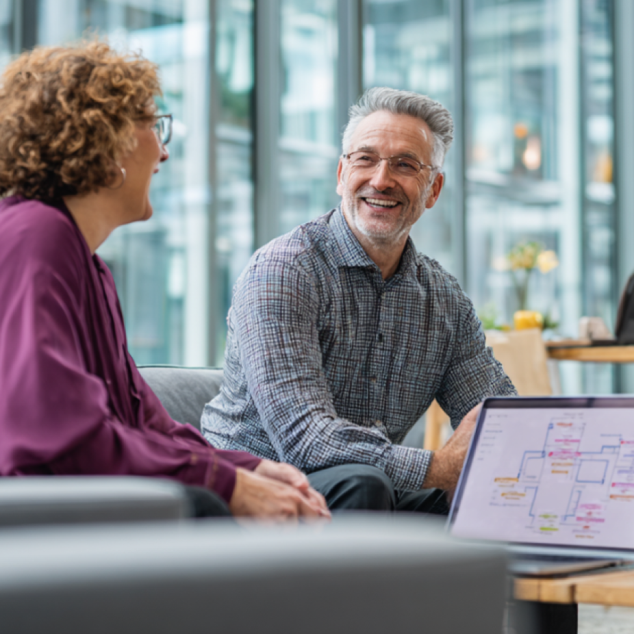 Two professionals have a relaxed conversation in a modern office lounge, with a laptop open beside them displaying a PPC campaign strategy map—symbolizing a discussion around paid search advertising strategies.