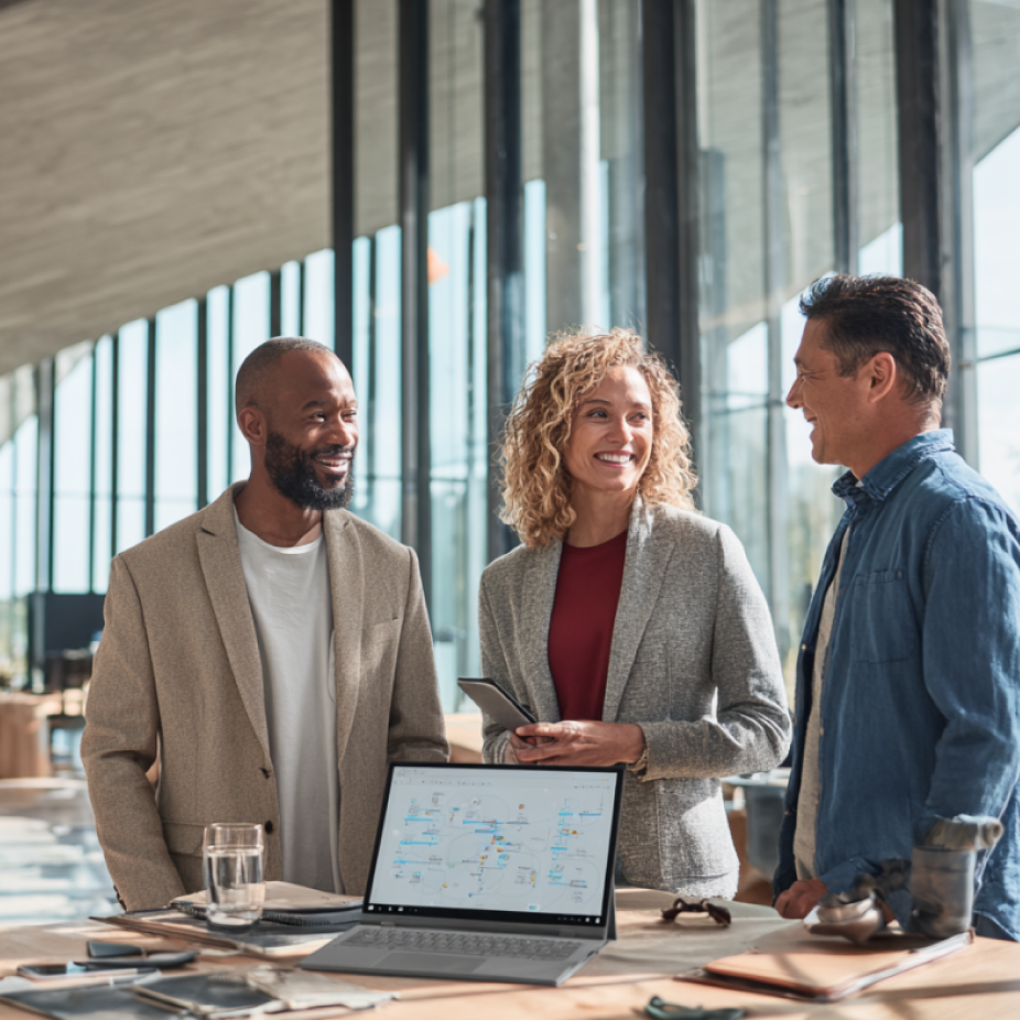 Three marketing professionals engaged in a collaborative discussion around a table in a bright modern office, with a laptop displaying a digital advertising strategy—representing social media advertising and campaign planning.