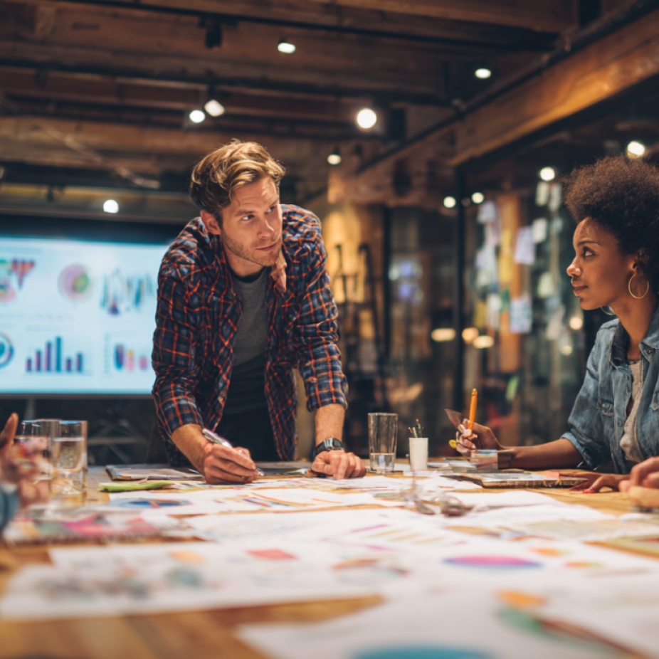 A team of marketing professionals collaborates around a table covered with colorful data charts and strategy papers, with digital graphs displayed on a screen in the background—illustrating programmatic advertising strategy development.