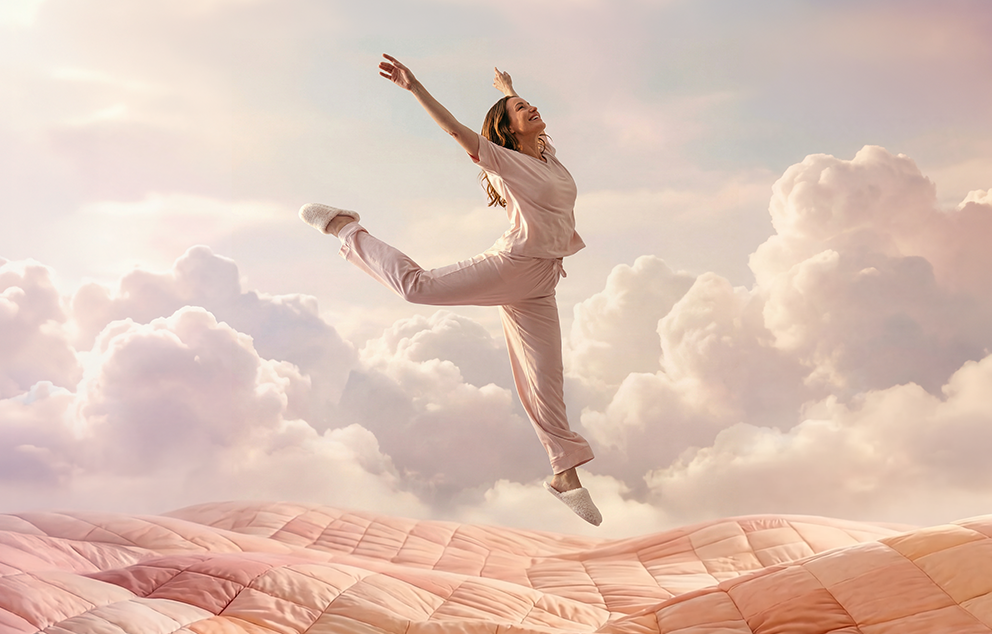 A woman jumps through a dream scene featuring pillows, blankets, and clouds as part of Keetsa’s “Designed to Dream On” brand concept.