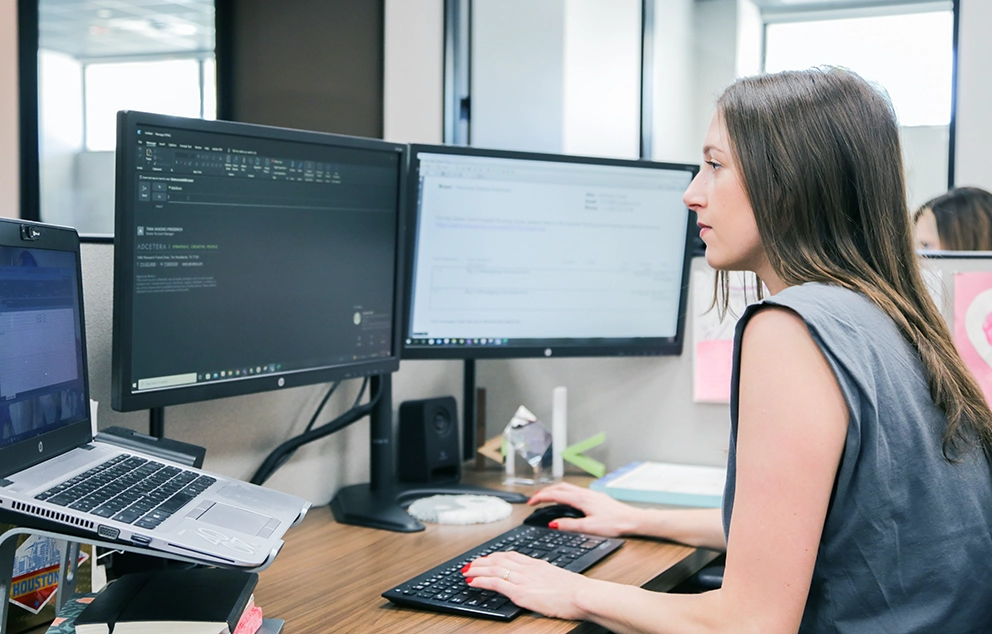 A woman sits at an office workstation with dual monitors and a laptop.