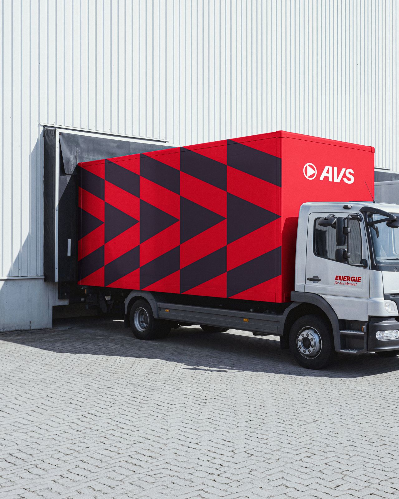 Roter AVS-Lieferwagen mit schwarzem Dreieckmuster lädt an Lagerhalle mit weißer Außenwand.