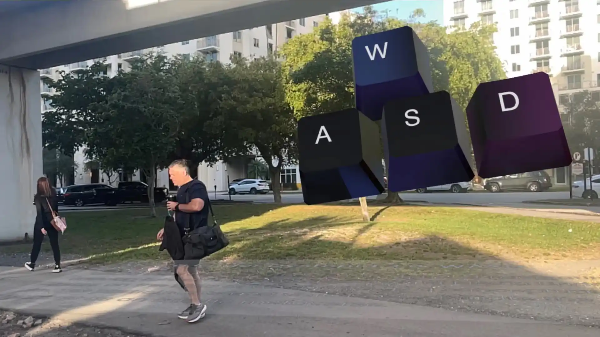 MUD Foundation WASD Keys Street Activation featuring large 3D W, A, S, and D keyboard keys floating above a grassy urban landscape, with a man walking while carrying a drink and a bag and a woman walking away nearby.