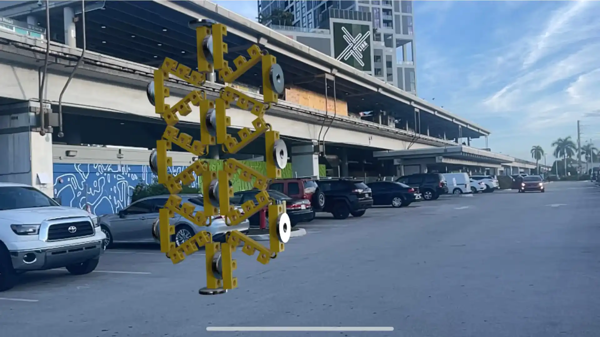 MUD Foundation Rafael Rodriguez’s Street Activation AR, featuring a large yellow geometric sculpture with interconnected angular forms and circular metal elements installed in an urban parking area with cars and modern buildings in the background.