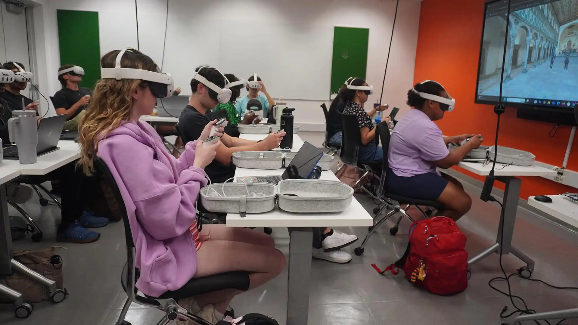 MUD Foundation XRCamp Spring 2026, featuring students wearing virtual reality headsets and holding VR controllers at desks with laptops and VR equipment, facing a large screen displaying a virtual corridor.