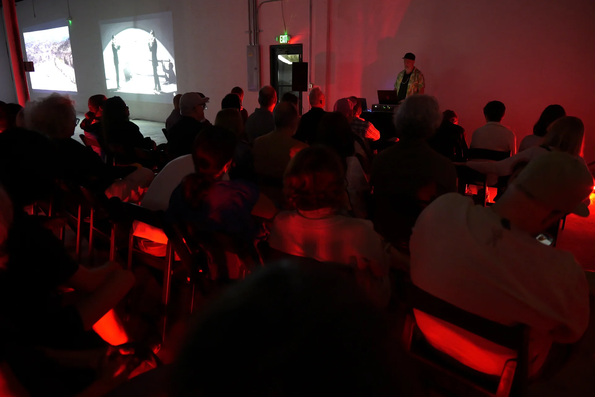 MUD Foundation The Sound of Art, featuring an audience seated in a dark exhibition space illuminated by red light while a performer operates electronic equipment, with projected video artworks displayed on the walls.