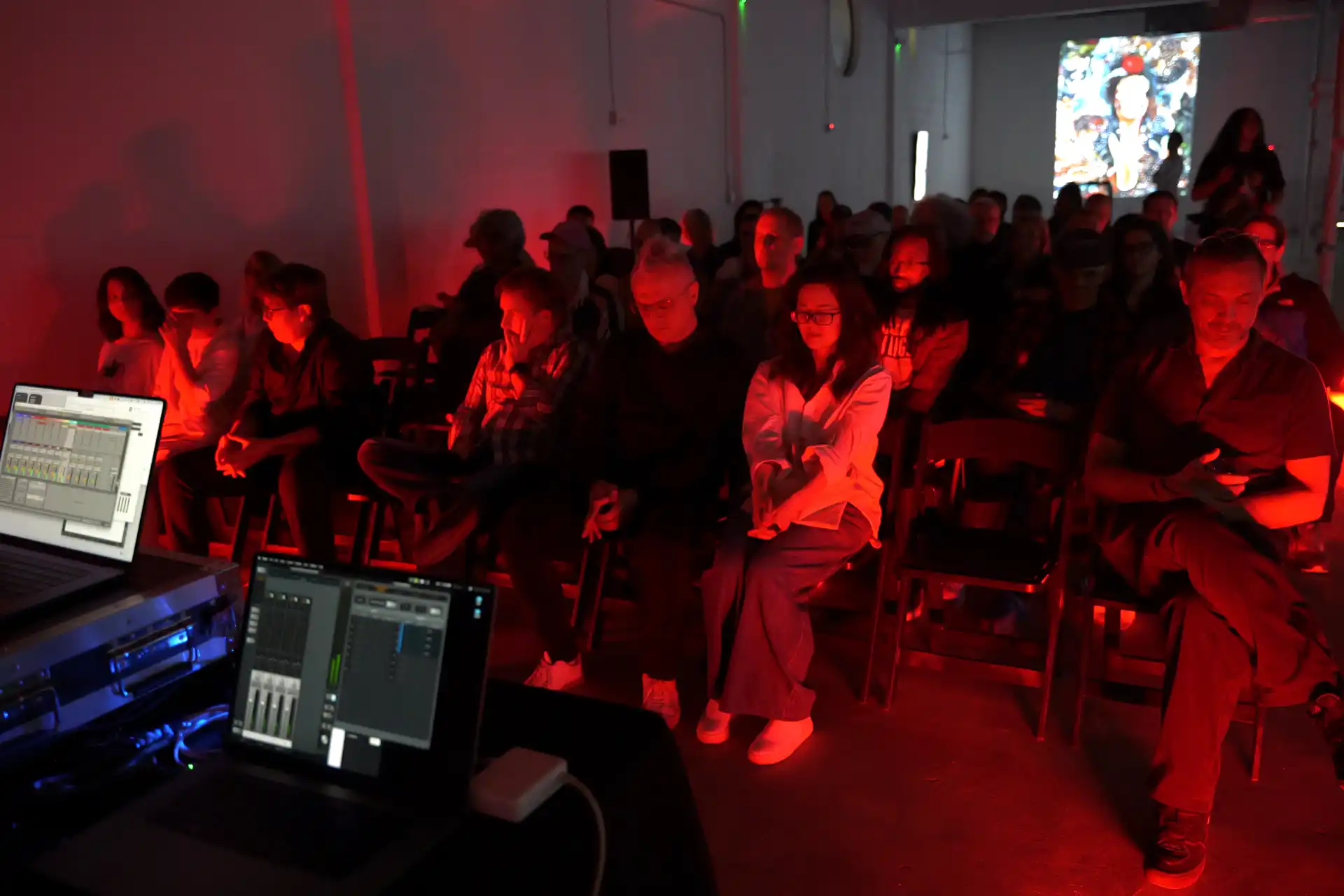 MUD Foundation The Sound of Art, featuring an audience seated in a dark exhibition space illuminated by red light, with audio equipment and mixing software visible in the foreground and a projected artwork in the background.