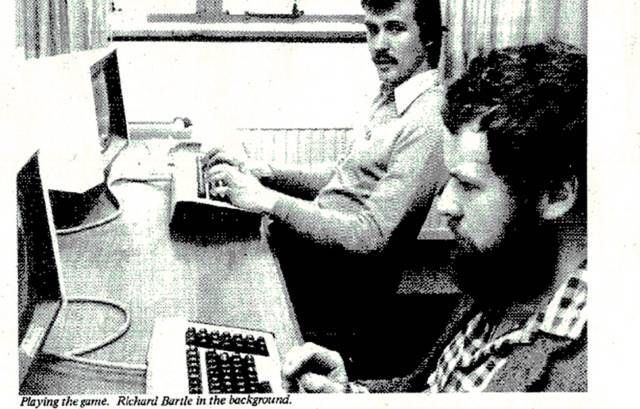 MUD Foundation archival photograph showing two men playing a computer game at desktop terminals, with Richard Bartle identified in the background.
