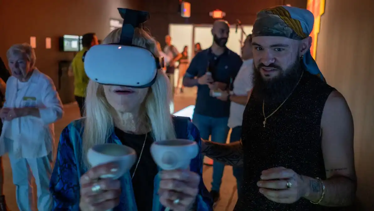 MUD Foundation Media Under Dystopia 3.0 exhibition program, featuring a woman wearing a VR headset and holding motion controllers while interacting with a virtual reality experience beside a man in a themed costume, with other visitors in the background.