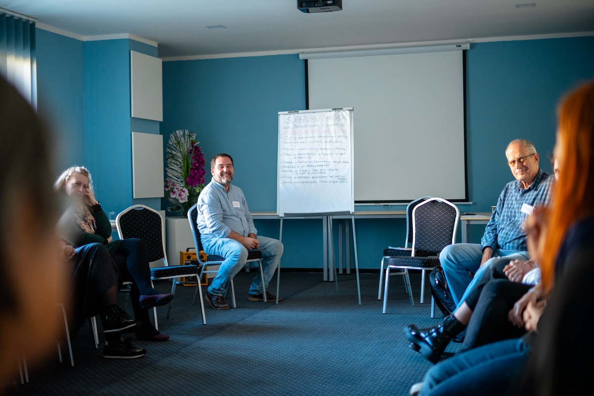 Skupina dospělých sedících v kruhu v místnosti na semináři nebo schůzce s modrou stěnou a tabulí v pozadí.