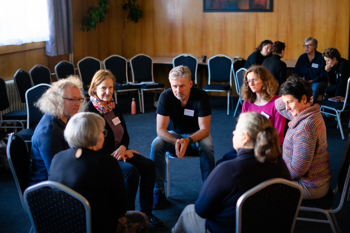 Skupina dospělých sedí v kruhu na židlích v místnosti s dřevěnými stěnami během diskuse.