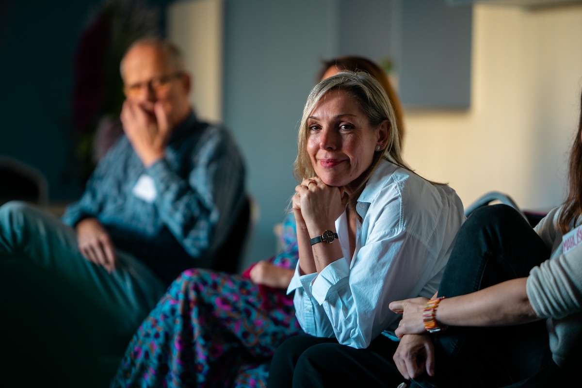 Žena v bílé košili sedí a usmívá se během neformální skupinové diskuse.