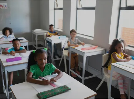 A classroom of children with a girl in a green shirt writing on a piece of paper.