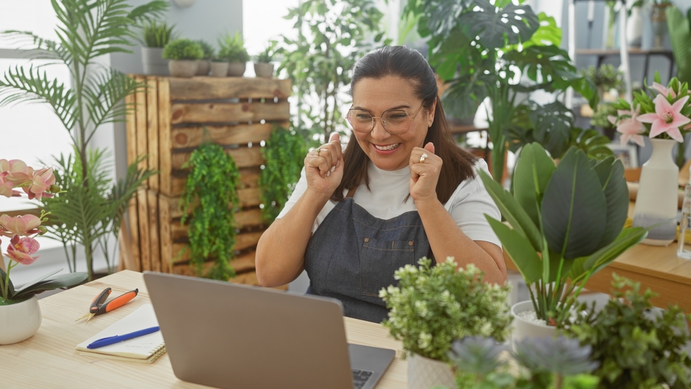Woman celebrating success while working on a laptop in a greenery-filled workspace, reflecting the importance of local SEO in enhancing business visibility.
