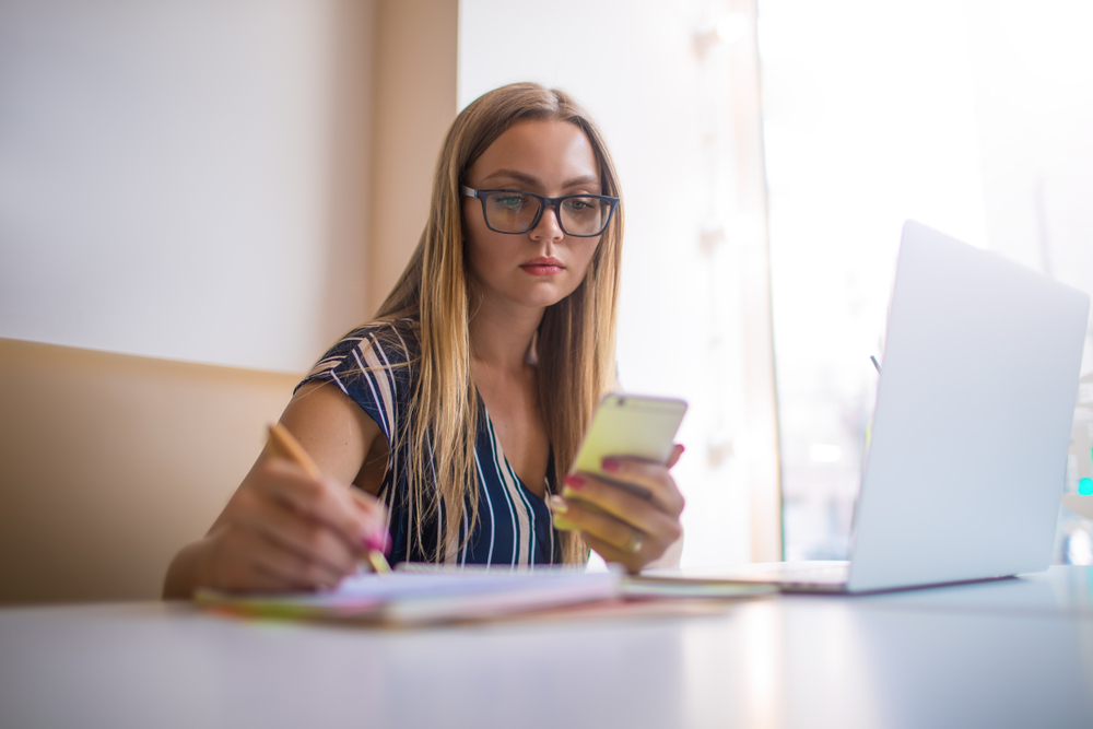 Woman using smartphone and taking notes at a table, emphasizing online research and local SEO strategies for small businesses.