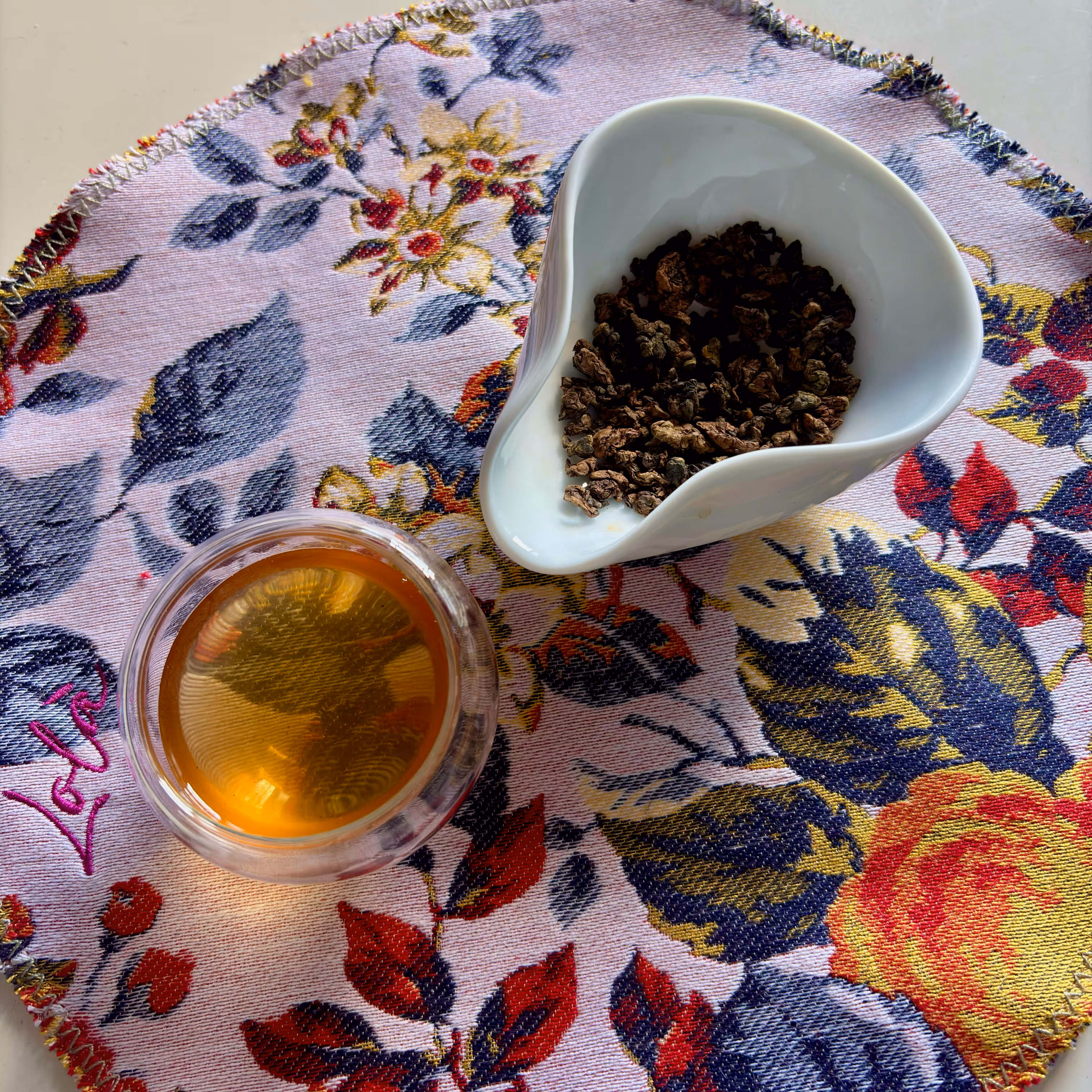Tazza di tè e ciotola con foglie di tè su tovaglietta con motivo floreale colorato.