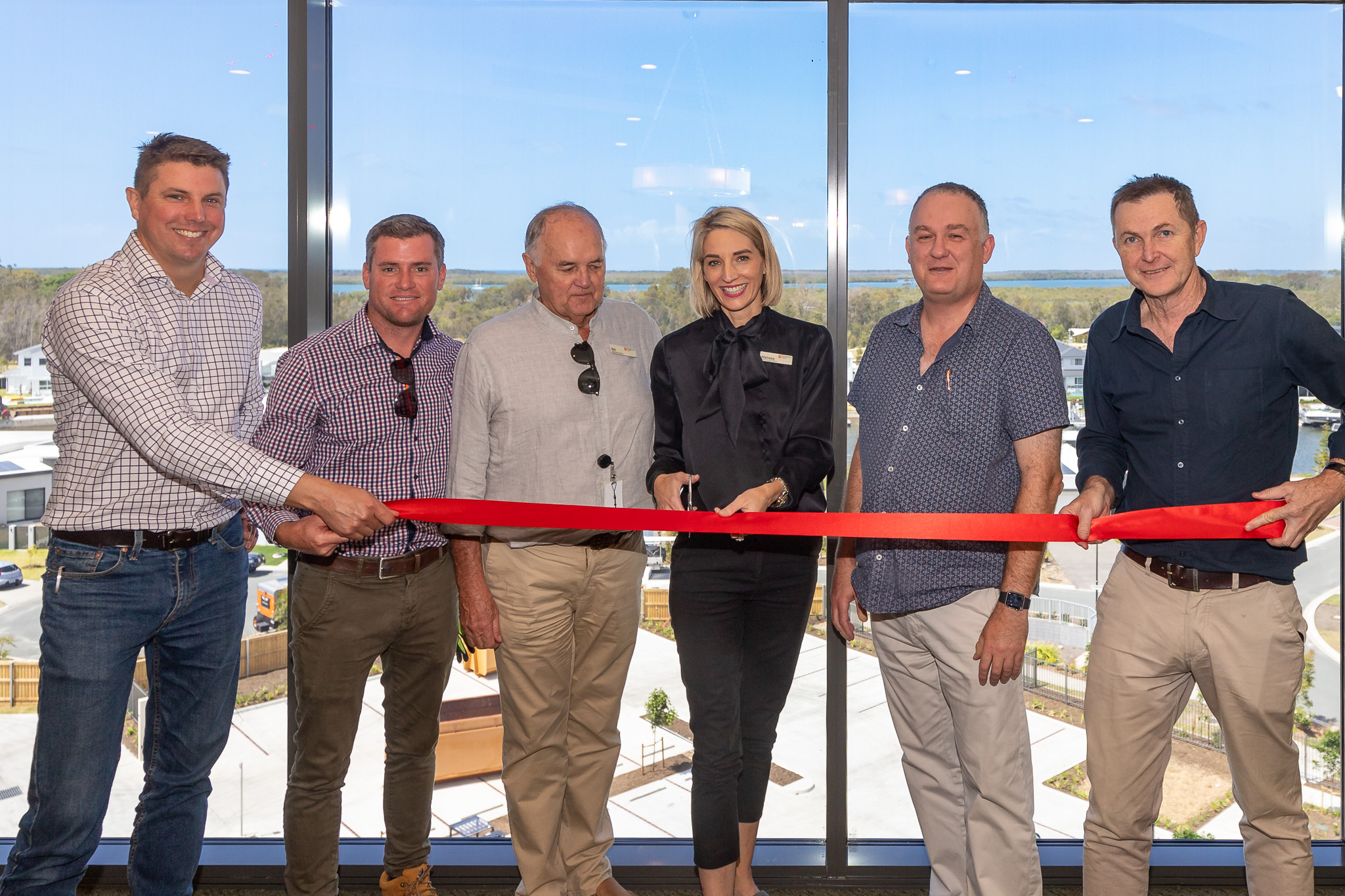 Six people standing in front of large windows cutting a red ribbon at a ribbon-cutting ceremony.