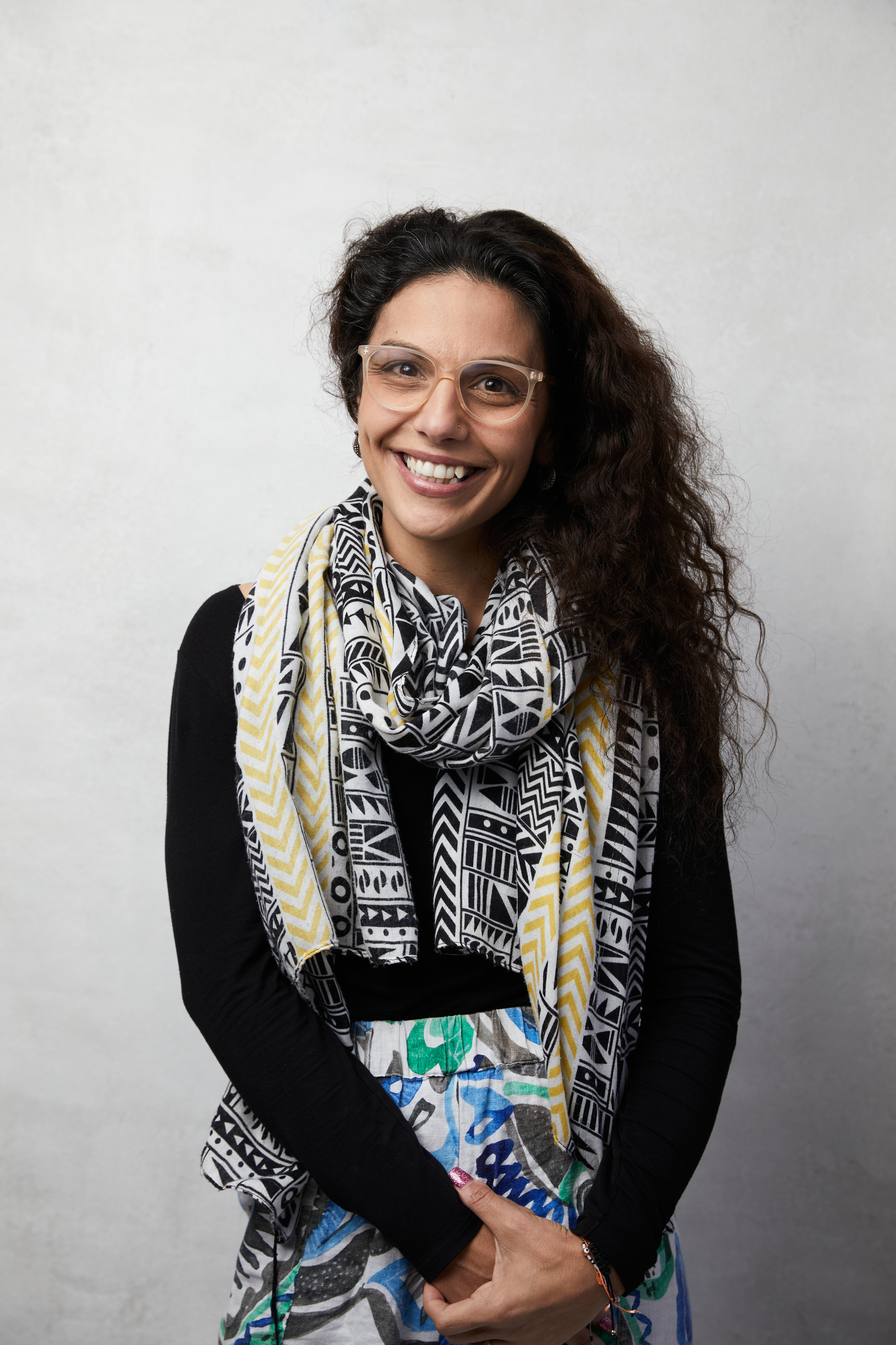 Smiling woman with curly dark hair wearing clear glasses, a black long-sleeve shirt, a patterned black and yellow scarf, and colorful patterned pants.