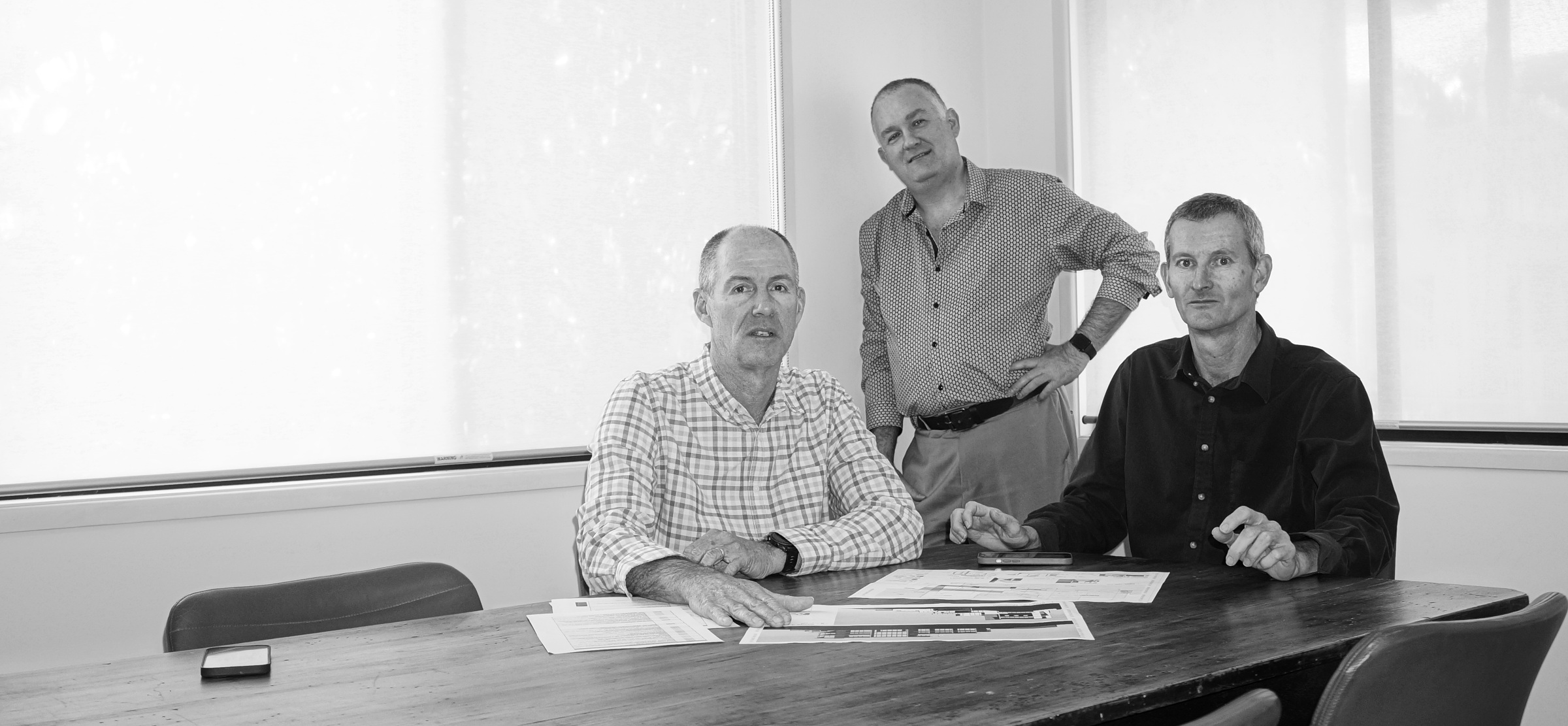 Three men in a meeting room with documents on a wooden table, two seated and one standing.